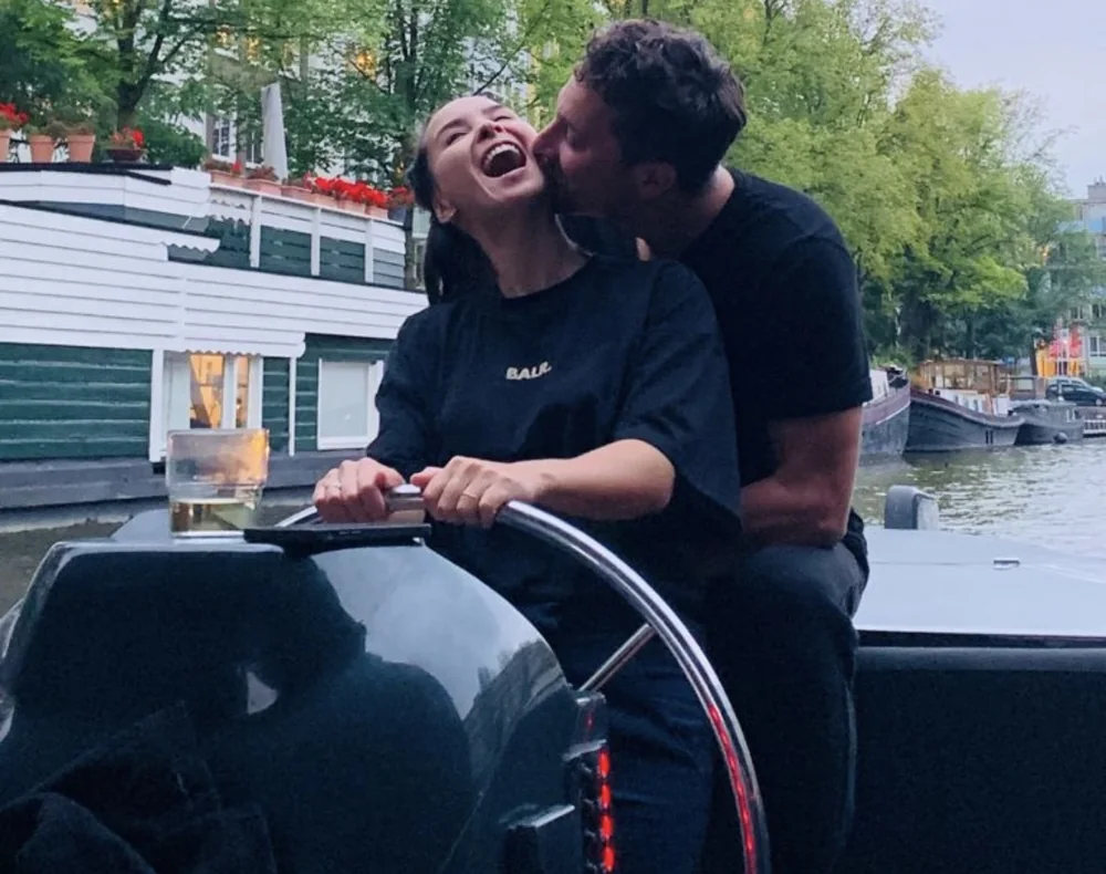 Eline aan het roer van een boot, hard lachend; Marc kust haar wang.