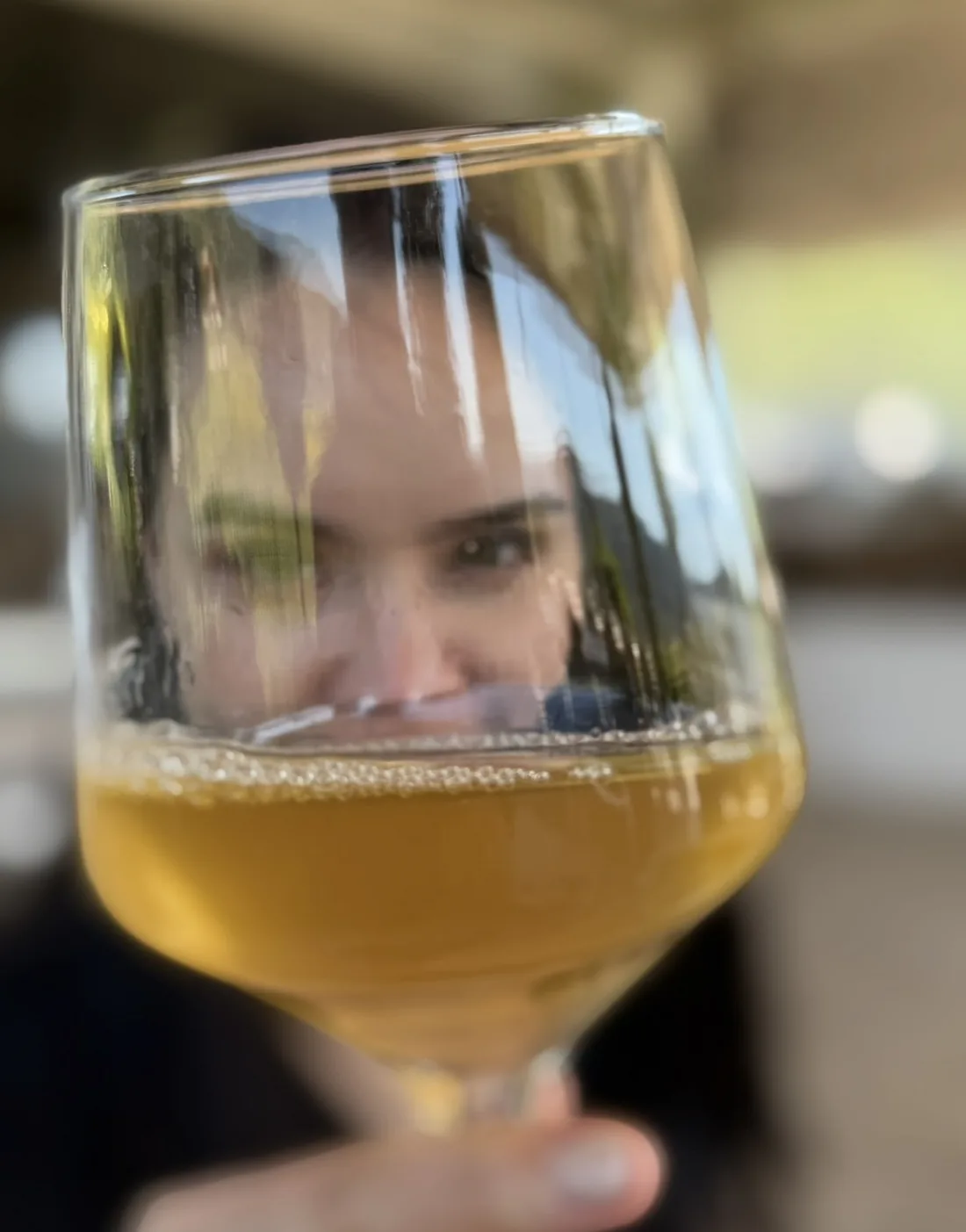 Eline Klaver kijkt door een wijnglas heen in de camera; haar gezicht is door het glas vervormd.