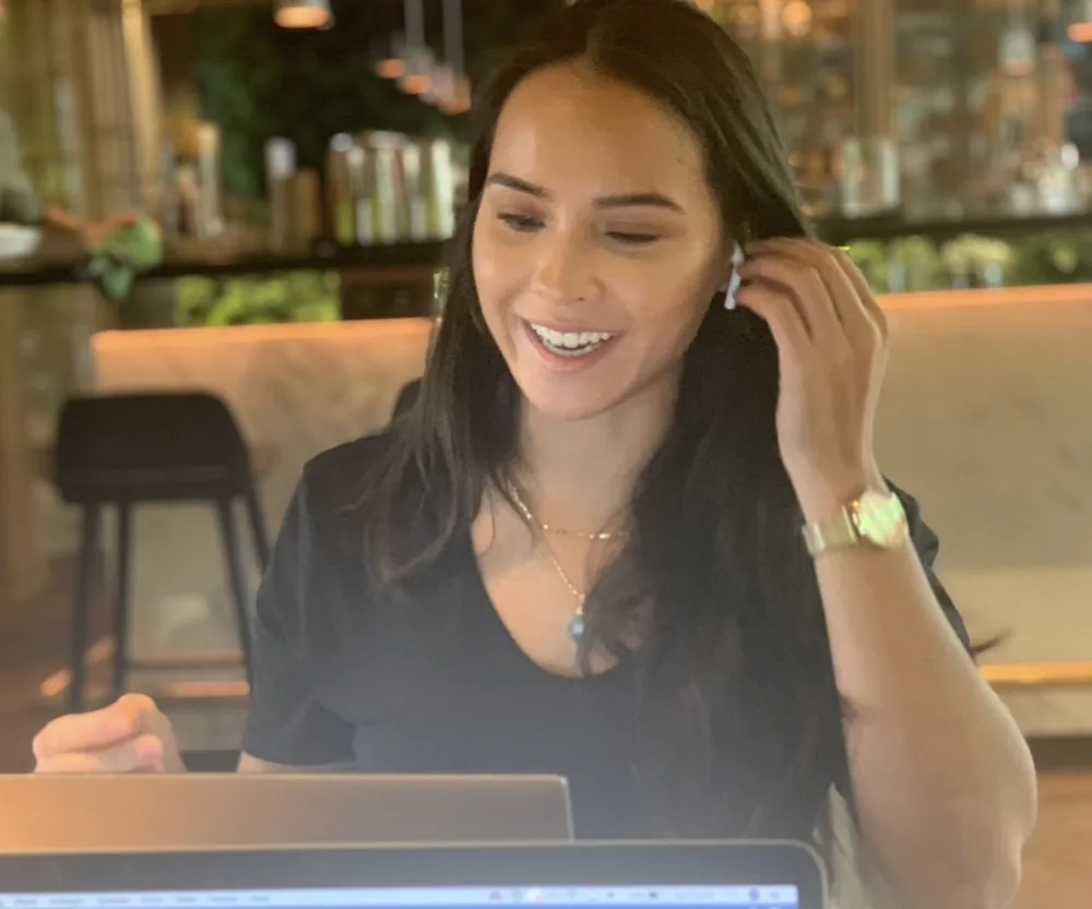 Eline lacht achter een laptop in een lounge, oortje in.