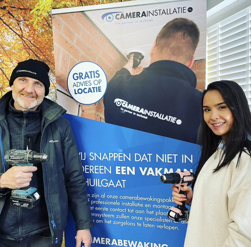 Eline met een roze boormachine naast een collega met een blauwe, voor een banner van het bedrijf.