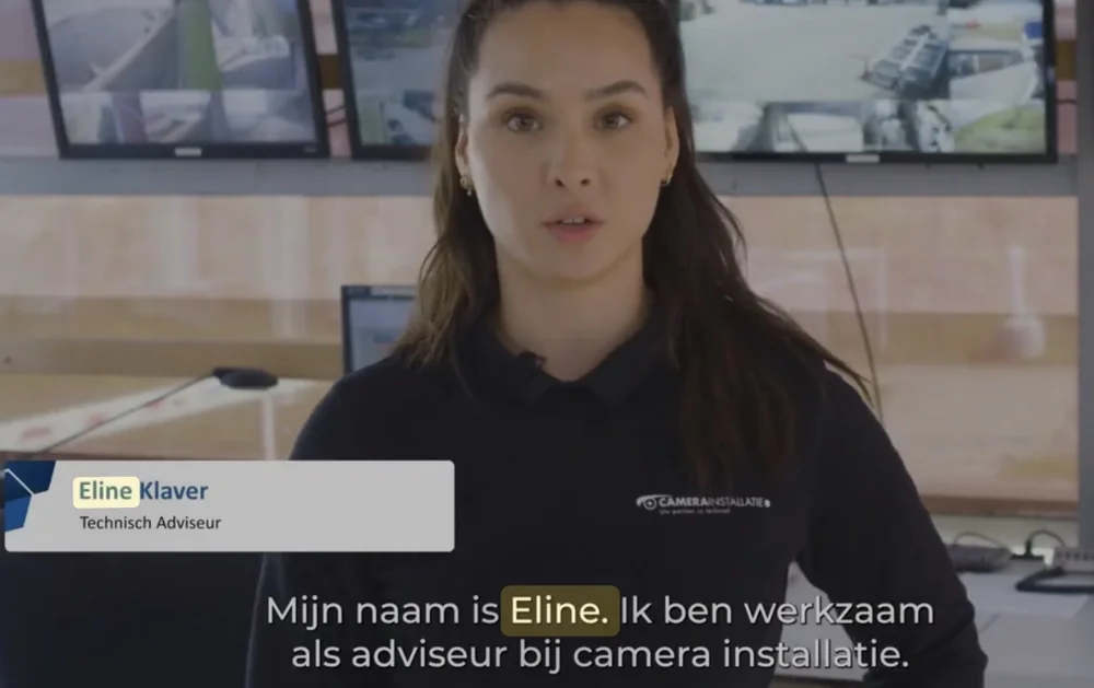 Videostill: Eline voor een wand met camerabeelden, met in beeld haar naam en functie, Technisch Adviseur, en de ondertitel: Mijn naam is Eline. Ik ben werkzaam als adviseur bij camera installatie.