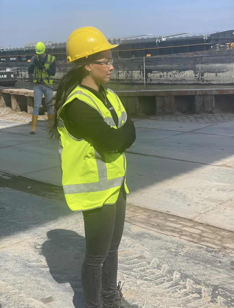 Eline op een werf, gele helm en geel hesje, armen over elkaar.